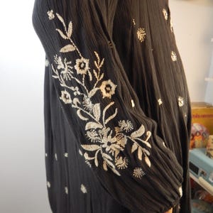 May include: Black long-sleeved dress with intricate white floral embroidery on the sleeves and scattered across the fabric. The dress has a textured appearance and a relaxed fit, suitable for casual wear.
