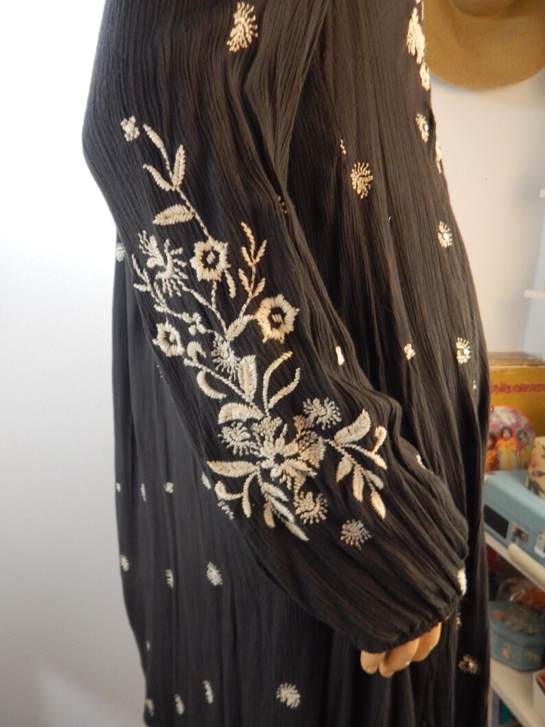 May include: Black long-sleeved dress with intricate white floral embroidery on the sleeves and scattered across the fabric. The dress has a textured appearance and a relaxed fit, suitable for casual wear.