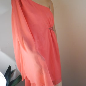 Puede incluir: Un vestido color coral con un solo hombro y un dise&ntilde;o fluido y drapeado. El vestido presenta un adorno decorativo en la cintura y un dobladillo con volantes en capas. La tela parece ligera y semitransparente, ideal para ocasiones formales.