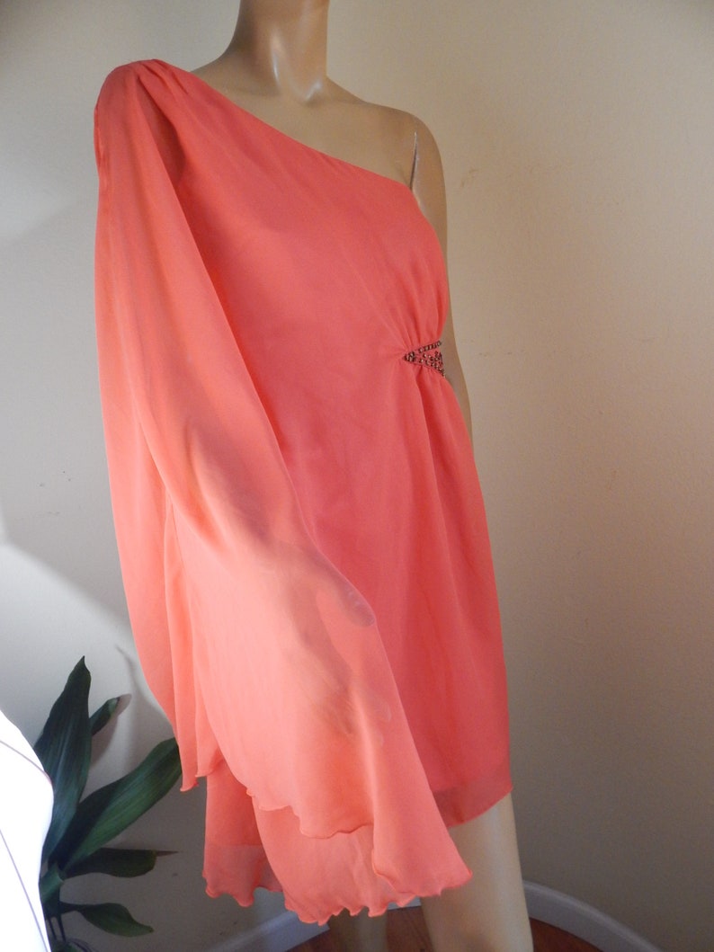 Puede incluir: Un vestido color coral con un solo hombro y un dise&ntilde;o fluido y drapeado. El vestido presenta un adorno decorativo en la cintura y un dobladillo con volantes en capas. La tela parece ligera y semitransparente, ideal para ocasiones formales.