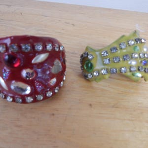 May include: Two decorative jewelry pieces. One is a wide, red cuff bracelet adorned with clear and red rhinestones and silver-colored accents. The other is a yellow-green, fan-shaped brooch with clear, green, and iridescent rhinestones.