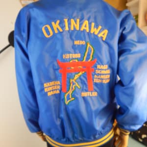 May include: Blue satin jacket with yellow and white trim. The back features the word "OKINAWA" in yellow lettering, along with a map and place names. The jacket has a classic bomber style with a collar and ribbed cuffs.