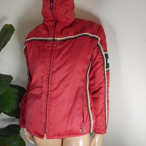 May include: A red jacket with a high collar and a full-length zipper. The jacket has white, black, and gold stripes on the sleeves and along the zipper. The jacket is displayed on a mannequin.