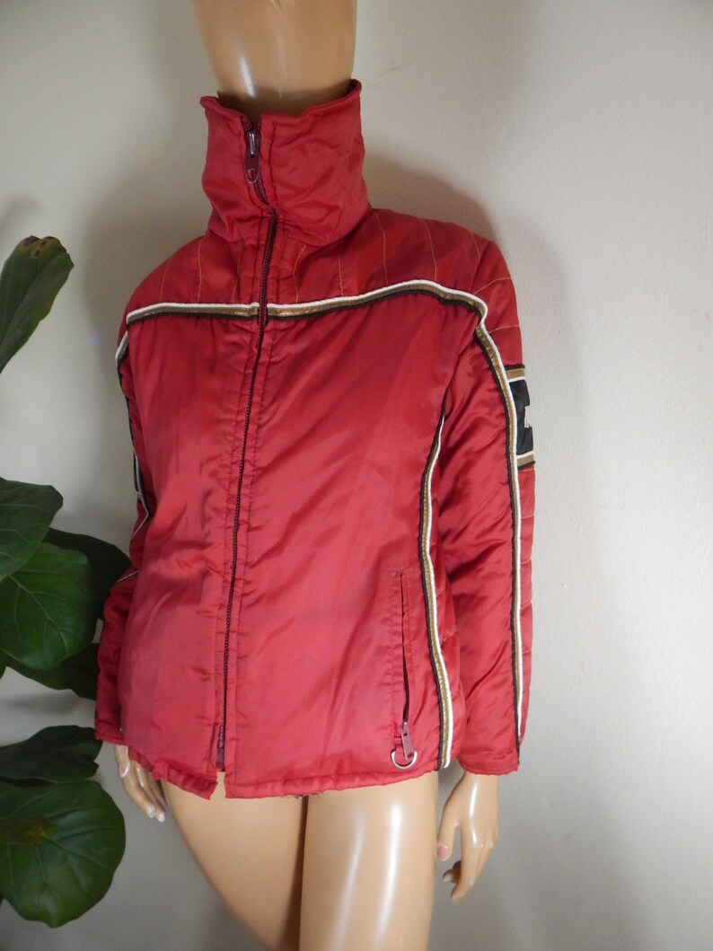 May include: A red jacket with a high collar and a full-length zipper. The jacket has white, black, and gold stripes on the sleeves and along the zipper. The jacket is displayed on a mannequin.