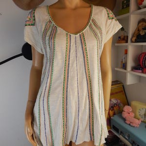 May include: Cream-colored short-sleeve top with a V-neckline. The shirt features vertical stripes of colorful embroidery in green, blue, and orange. The sleeves and neckline are also adorned with embroidered details. The top has a slightly scalloped hemline.