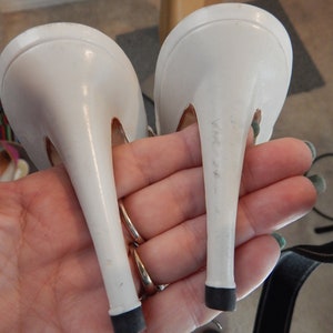 May include: A pair of white stiletto high-heeled shoes. The heels are slender and have black rubber tips. The shoes are held in a hand, showing the underside of the heels and the shoe's design.