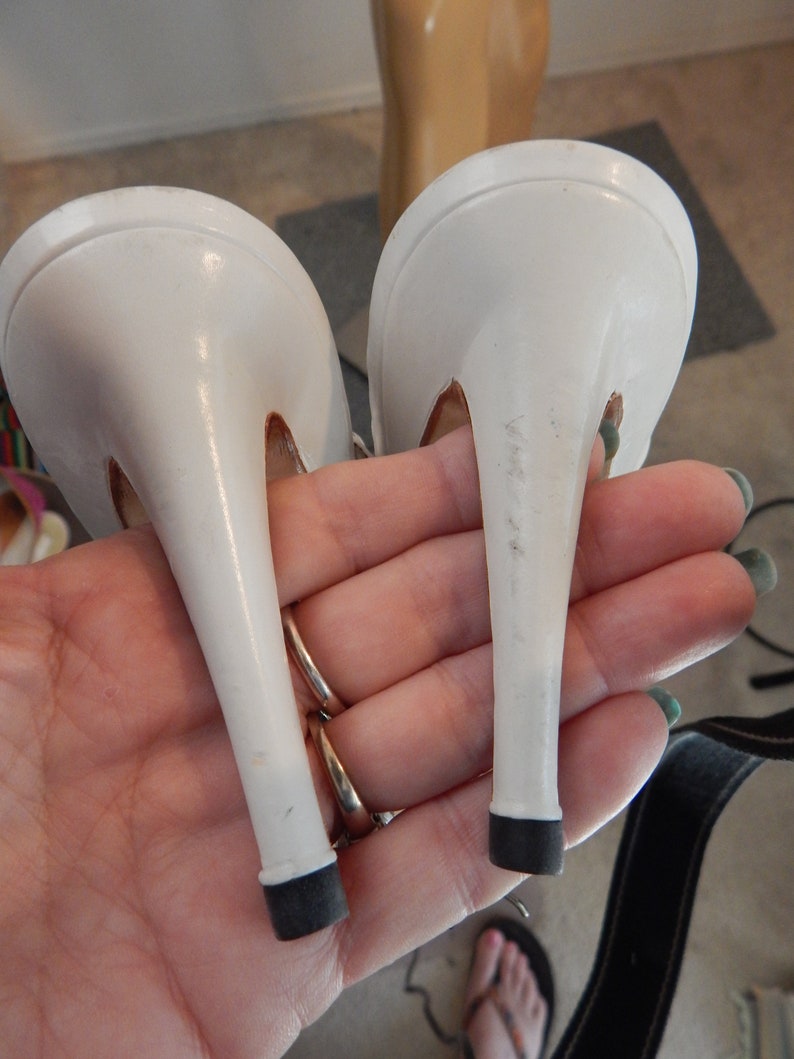May include: A pair of white stiletto high-heeled shoes. The heels are slender and have black rubber tips. The shoes are held in a hand, showing the underside of the heels and the shoe's design.