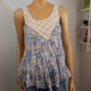 May include: A sleeveless top with a floral pattern in shades of blue and cream. The top features a cream-colored, open-weave crochet panel at the neckline. The bottom of the top has a ruffled hemline. The top is displayed on a mannequin.