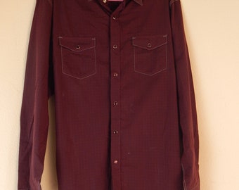 May include: A long-sleeved, burgundy button-down shirt with two chest pockets and white stitching. The shirt has a classic collar and is hanging on a pink hanger.