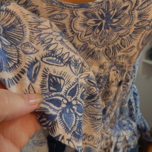 May include: A light-colored top with a blue floral pattern. The fabric is soft and the design features detailed, stylized flowers and leaves. The top has a gathered waist and ruffled sleeves, creating a flowing silhouette.