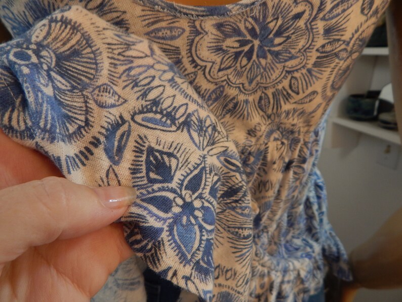 May include: A light-colored top with a blue floral pattern. The fabric is soft and the design features detailed, stylized flowers and leaves. The top has a gathered waist and ruffled sleeves, creating a flowing silhouette.