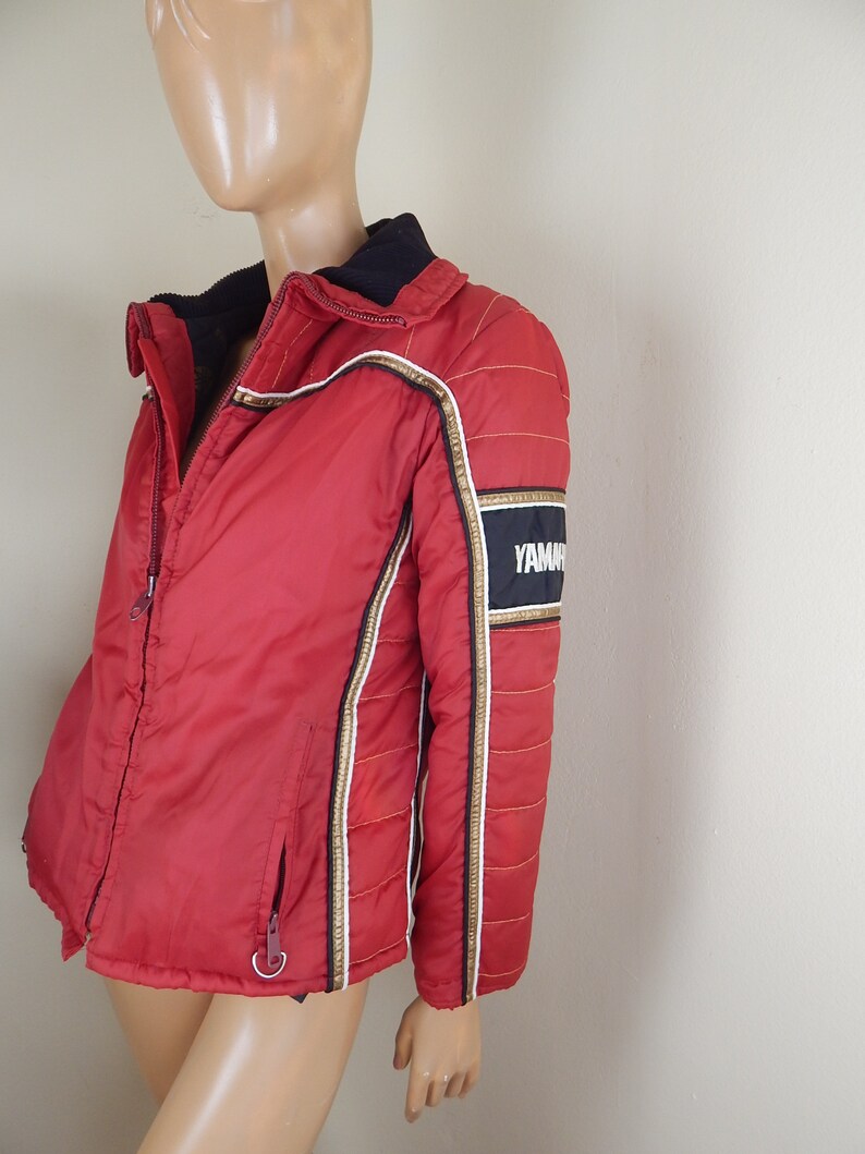 May include: Red and black Yamaha racing jacket with gold and white accents. The jacket has a black collar and a zipper closure. The Yamaha logo is displayed on the sleeve.