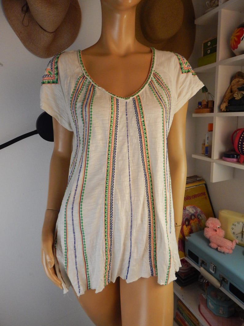 May include: Cream-colored short-sleeve top with a V-neckline. The shirt features vertical stripes of colorful embroidery in green, blue, and orange. The sleeves and neckline are also adorned with embroidered details. The top has a slightly scalloped hemline.