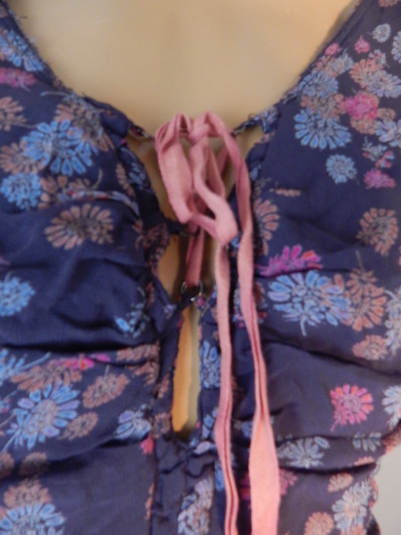 May include: A dark blue floral top with a V-neckline and a pink ribbon tie. The fabric is patterned with blue, pink, and peach-colored flowers. The top has a gathered design.