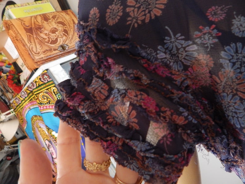 May include: Dark navy blue sheer fabric with a floral pattern in shades of pink, orange, and blue. The edge of the fabric has a ruffled trim. The fabric is lightweight and appears to be a blouse or dress material.