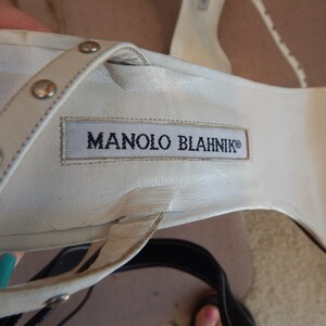 May include: Close-up of a white leather sandal with silver studs on the straps. The sandal features the brand name "MANOLO BLAHNIK" printed on the insole. The shoe is a high-heeled sandal.
