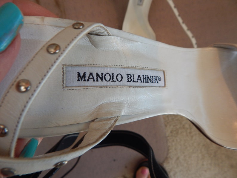 May include: Close-up of a white leather sandal with silver studs on the straps. The sandal features the brand name "MANOLO BLAHNIK" printed on the insole. The shoe is a high-heeled sandal.
