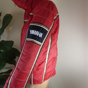 May include: Red Yamaha racing jacket with gold and white accents. The jacket features a black and gold patch with the Yamaha logo on the sleeve. The jacket has a quilted design and a high collar.