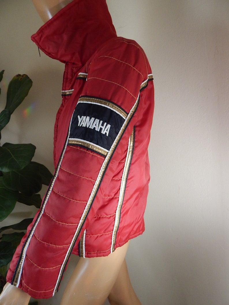 May include: Red Yamaha racing jacket with gold and white accents. The jacket features a black and gold patch with the Yamaha logo on the sleeve. The jacket has a quilted design and a high collar.