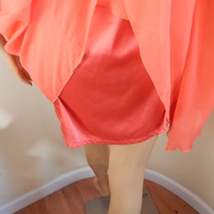 Puede incluir: Conjunto de lencer&iacute;a color coral. El conjunto incluye una enagua de sat&eacute;n y una sobrecapa transparente y fluida. La tela se drapea elegantemente, mostrando la textura y el color de la prenda. El estilo general sugiere una est&eacute;tica vintage o rom&aacute;ntica.