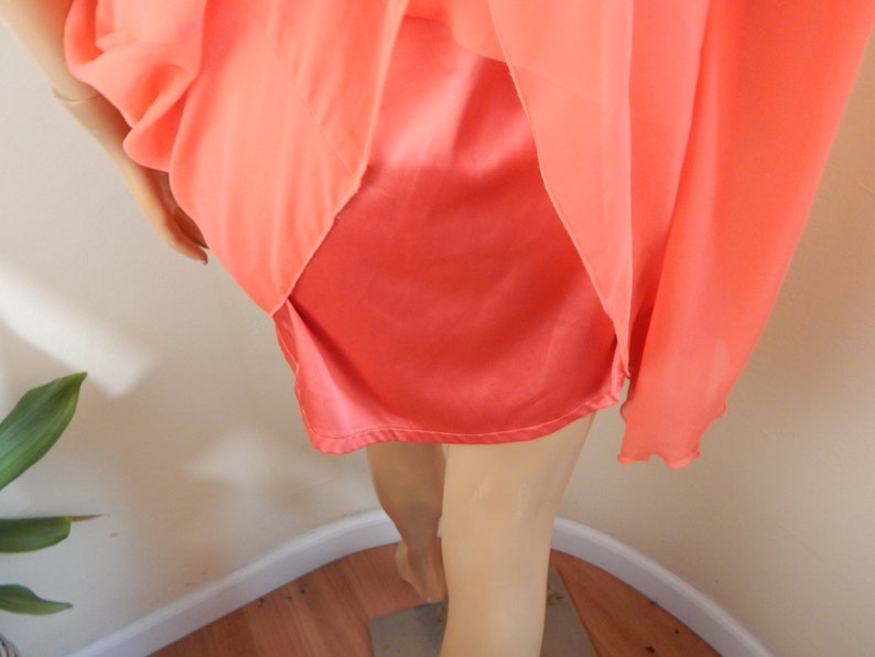 Puede incluir: Conjunto de lencer&iacute;a color coral. El conjunto incluye una enagua de sat&eacute;n y una sobrecapa transparente y fluida. La tela se drapea elegantemente, mostrando la textura y el color de la prenda. El estilo general sugiere una est&eacute;tica vintage o rom&aacute;ntica.