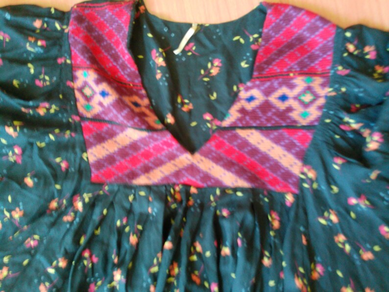 May include: Dark teal dress with floral print and a patterned yoke. The yoke features red, orange, and purple geometric designs. The dress has a round neckline and gathered skirt.
