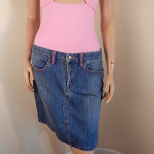 May include: A denim skirt with a classic five-pocket design. The skirt is a medium wash blue with a slight fade and features a zipper and button closure. The waistband has pink stitching. The skirt is paired with a pink bodysuit.