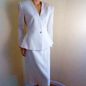 May include: Ivory-colored skirt suit. The jacket has a V-neckline with sequin and bead detailing, padded shoulders, and a single button closure. The skirt is knee-length. The suit is displayed on a mannequin.