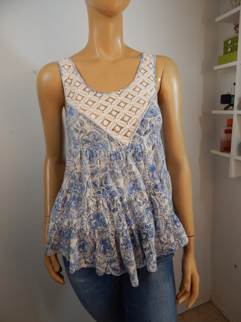 May include: A sleeveless top with a floral pattern in shades of blue and cream. The top features a cream-colored, open-weave crochet panel at the neckline. The bottom of the top has a ruffled hemline. The top is displayed on a mannequin.