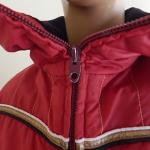 May include: A red winter jacket with a hood and a full zipper. The jacket has gold and white stripes across the chest and gold stitching. The collar is black.