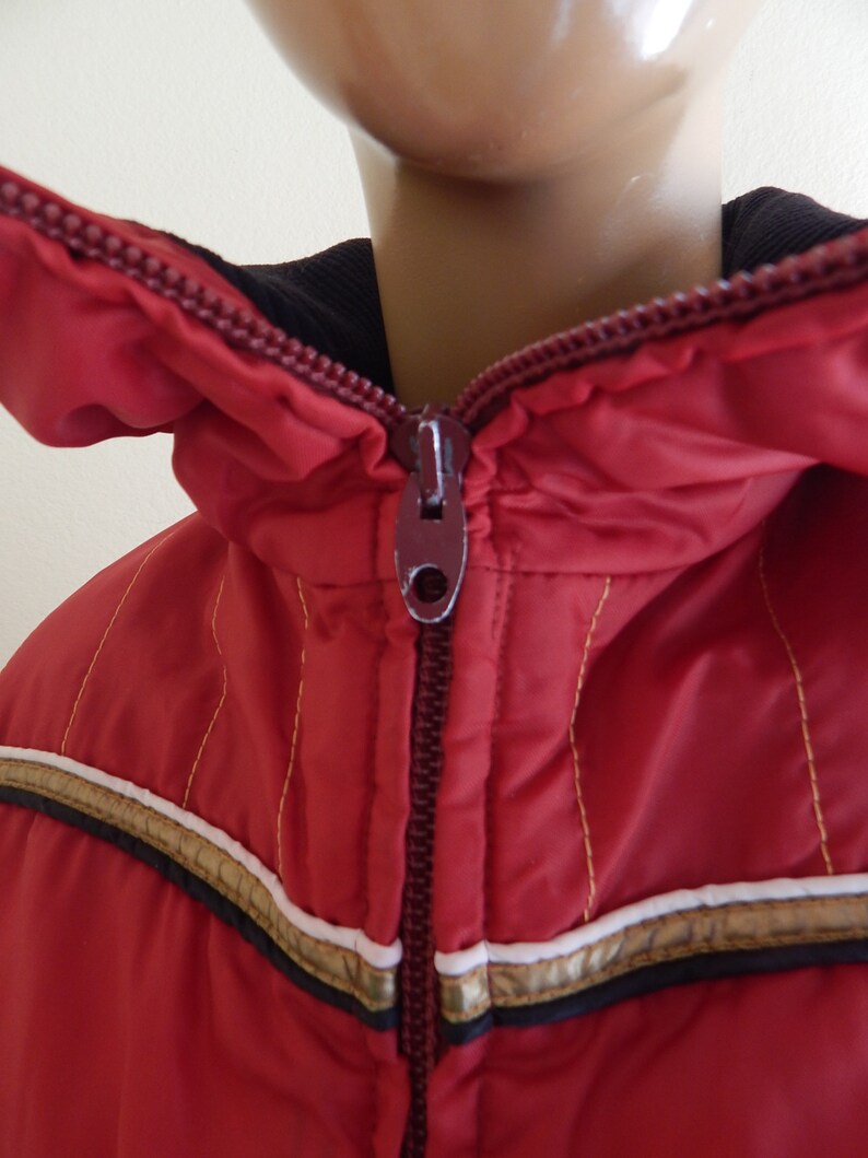 May include: A red winter jacket with a hood and a full zipper. The jacket has gold and white stripes across the chest and gold stitching. The collar is black.