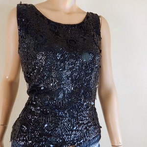 May include: A sleeveless black sequin top with a scoop neckline. The top is embellished with sequins and beaded fringe. The garment is displayed on a mannequin, paired with blue jeans. The top is a vintage style.