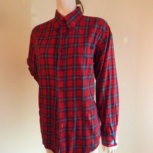 May include: A red plaid button-down shirt with long sleeves, displayed on a mannequin. The plaid pattern includes teal and navy blue lines. The shirt is buttoned up and has a collar and a chest pocket.