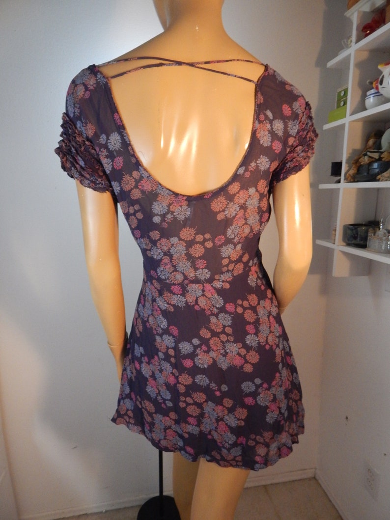 May include: A dark purple dress with a floral pattern in pink, blue, and peach. The dress features a low back with crisscross straps and short, ruffled sleeves. The dress is displayed on a mannequin.