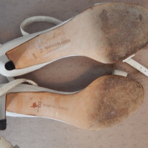 May include: Pair of white leather high-heeled sandals with ankle straps. The soles are worn and show the brand name "Manolo Blahnik" and the size "39 1/2". The heels are approximately 7.6 cm high.