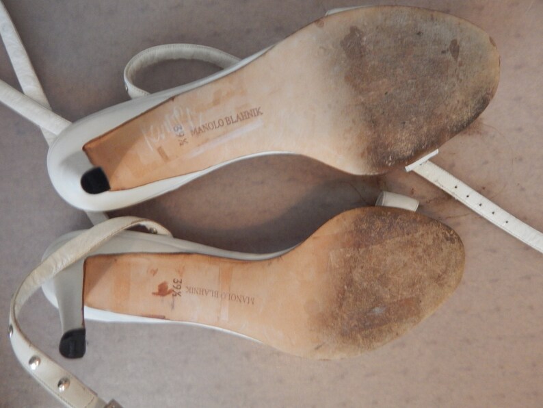May include: Pair of white leather high-heeled sandals with ankle straps. The soles are worn and show the brand name "Manolo Blahnik" and the size "39 1/2". The heels are approximately 7.6 cm high.