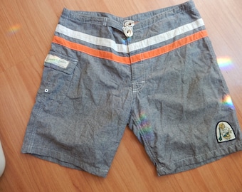 May include: Gray board shorts with white and orange stripes across the front. The shorts have a small pocket on the side and a patch with a cartoon character. The shorts have a drawstring closure.