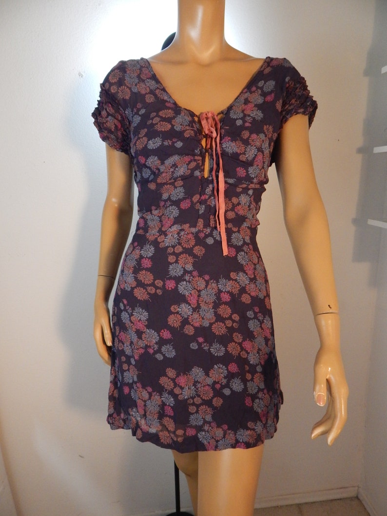 May include: A dark purple floral print dress with a V-neckline and short sleeves. The dress features a tie detail at the bust and a flared skirt. The floral pattern includes pink, orange, and blue flowers. The dress is displayed on a mannequin.