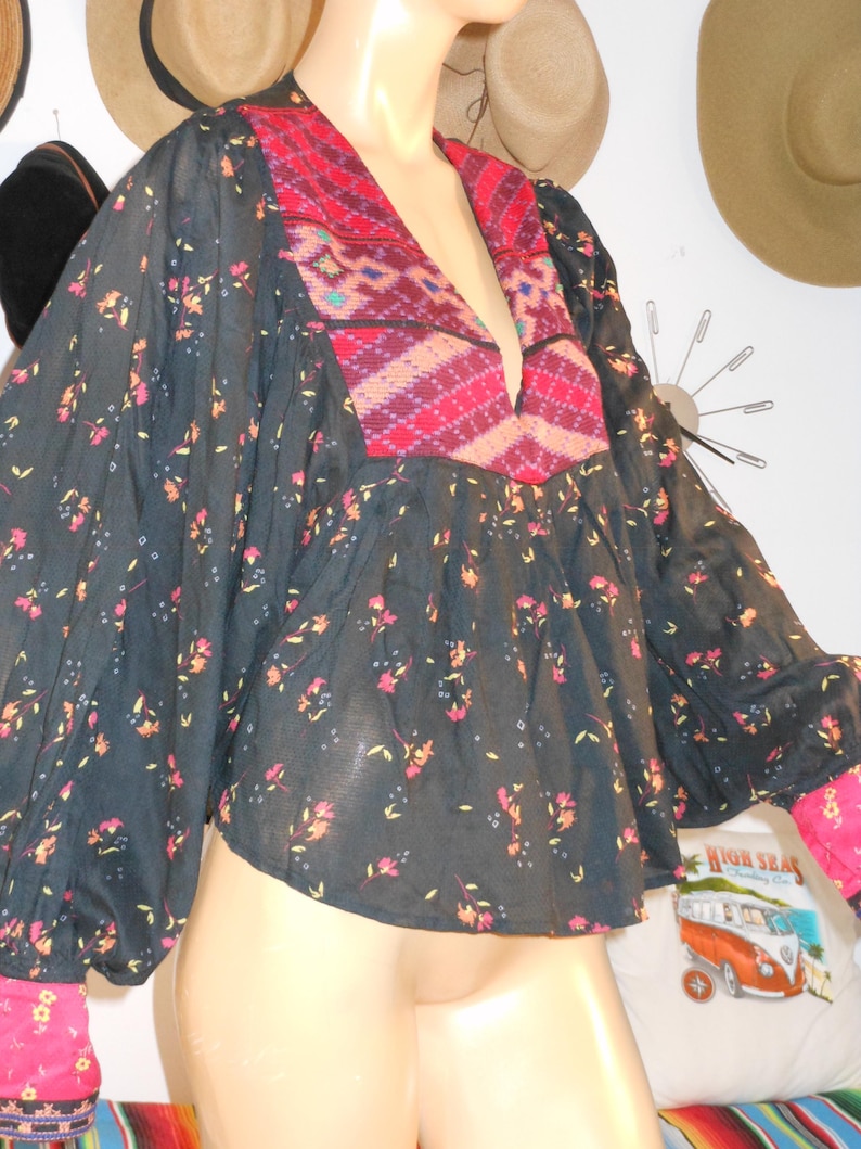 May include: A dark navy blouse with a floral pattern and a deep V-neck. The neckline and cuffs feature a vibrant red and pink geometric design. The sleeves are long and billowy, and the cuffs are a bright pink.
