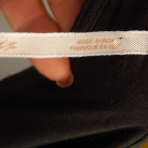 May include: Close-up of a white fabric tag with the text "MADE IN INDIA" and "FABRIQUE EN INDE" in brown lettering. The tag is attached to a dark gray garment. A small brown tag is visible.