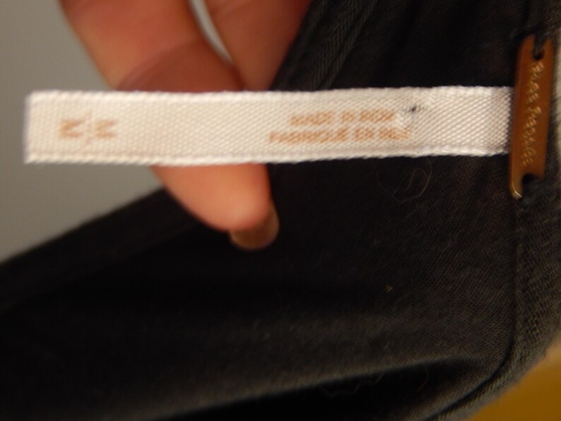 May include: Close-up of a white fabric tag with the text "MADE IN INDIA" and "FABRIQUE EN INDE" in brown lettering. The tag is attached to a dark gray garment. A small brown tag is visible.