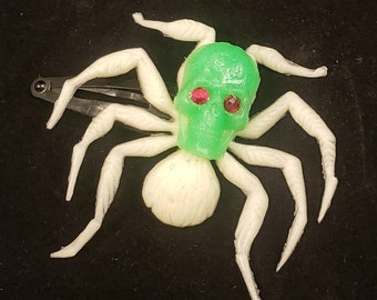 Puede incluir: Una araña de plástico blanca con una cabeza de calavera verde y ojos de gema roja. La araña tiene ocho patas y es una decoración de Halloween.