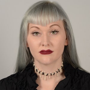 May include: A woman with silver hair and dark red lipstick is wearing a black choker necklace with white bone-like accents.