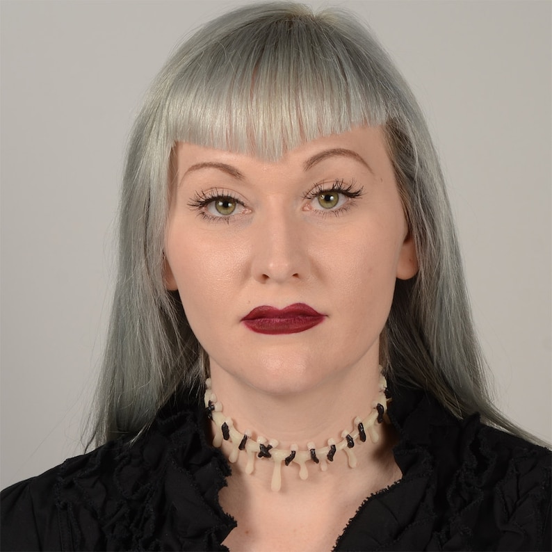 May include: A woman with silver hair and dark red lipstick is wearing a black choker necklace with white bone-like accents.