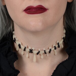 May include: A white and black choker necklace with dripping details, resembling stitches or bones. The necklace is worn by a person with dark red lipstick.