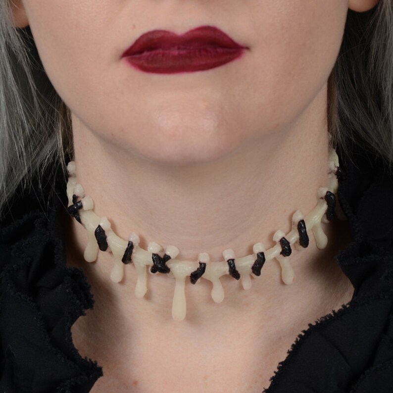 May include: A white and black choker necklace with dripping details, resembling stitches or bones. The necklace is worn by a person with dark red lipstick.