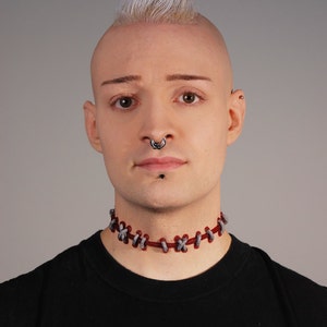 May include: A person with a shaved head and a mohawk hairstyle is wearing a black t-shirt and a red and gray choker necklace. They have a nose ring and a stud earring in their left ear.
