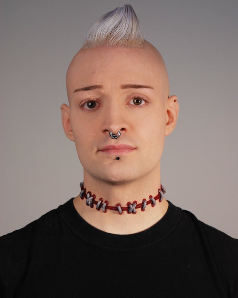 May include: A person with a shaved head and a mohawk hairstyle is wearing a black t-shirt and a red and gray choker necklace. They have a nose ring and a stud earring in their left ear.