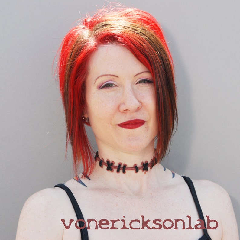May include: A woman with red and brown hair wearing a black choker with brown stitching. The choker is made of a braided material. The woman is wearing a black tank top.
