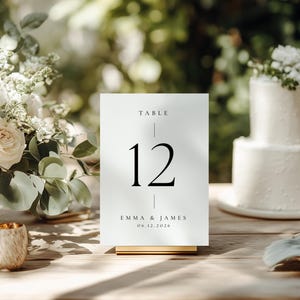 以下が含まれることがあります： TABLE 12、EMMA & JAMES、06.12.2026と黒い文字で書かれた長方形の白いテーブルナンバーサイン。ゴールドカラーのスタンドに立てられており、テキスト要素を区切る細い垂直線が特徴のミニマルなデザインです。
